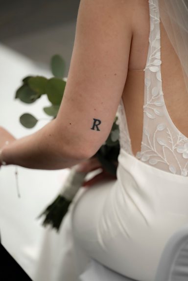 Braut mit 'R'-Tattoo auf dem linken Oberarm, trägt ein elegantes Hochzeitskleid und hält Blumen.